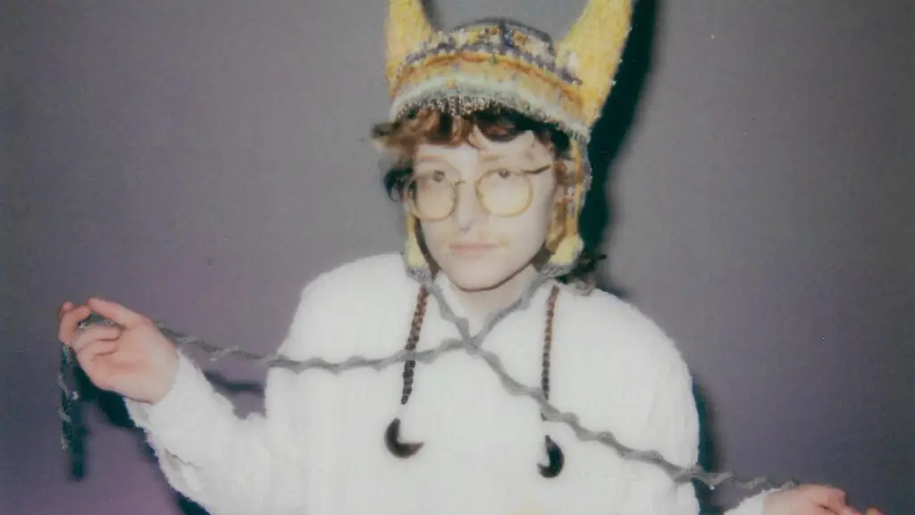 Cavetown in press photo wearing round glasses and yellow knit hat, holding yarn strings with crescent moon pendants