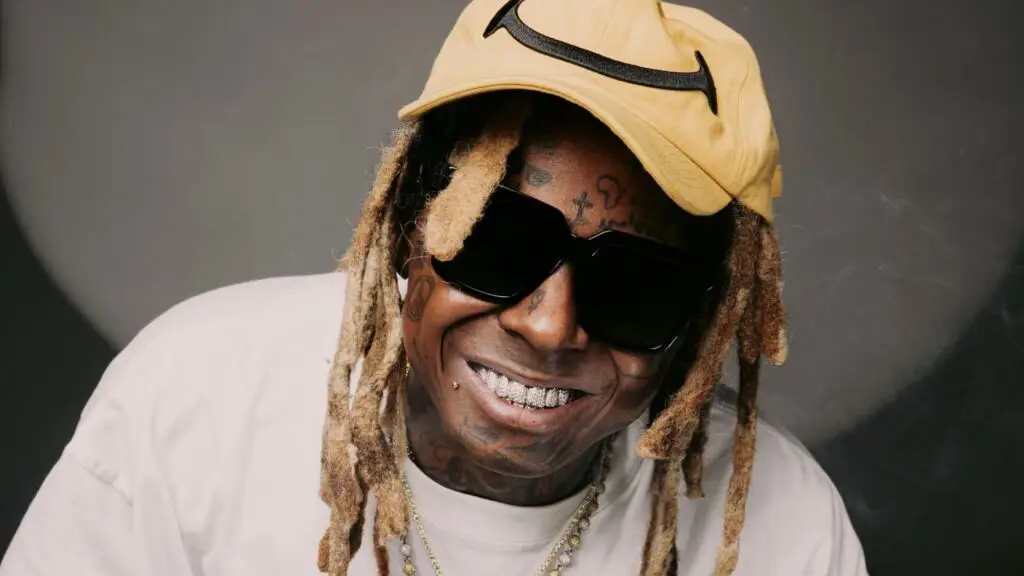 Lil Wayne smiling in promotional photo wearing yellow Nike cap, sunglasses, and signature dreadlocks