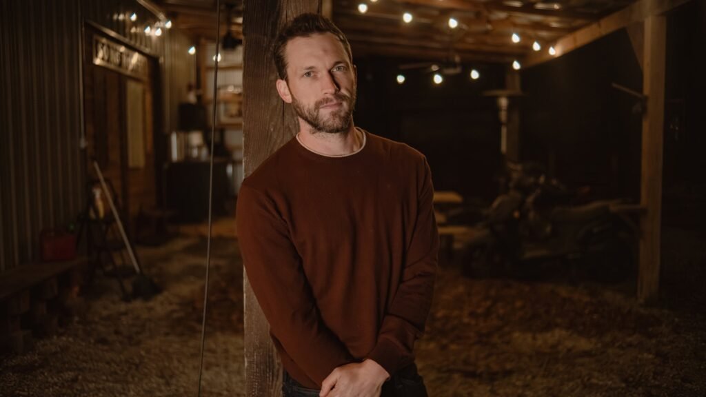 Phillip Phillips official press photo, leaning against a wooden post in a rustic barn with string lights