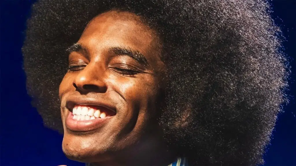 Akeem Ali performing live, smiling with eyes closed, large natural afro, blue stage lighting