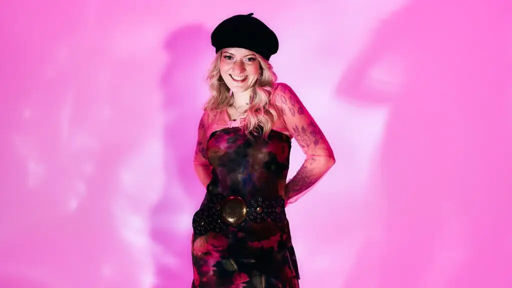 Scene Queen performing live, smiling against a pink backdrop in beret and floral dress