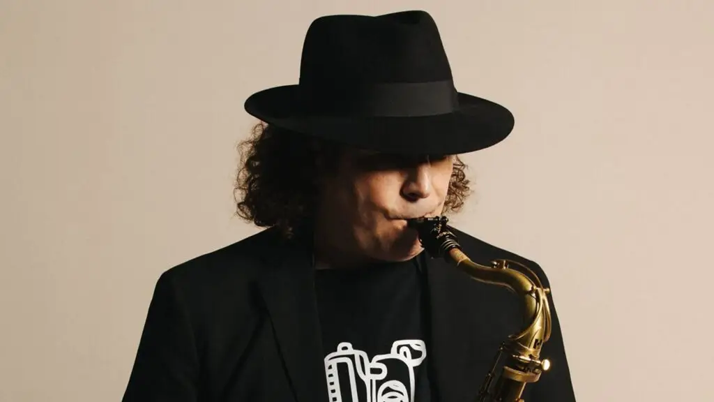 Boney James plays saxophone wearing a black fedora and blazer in official press photo