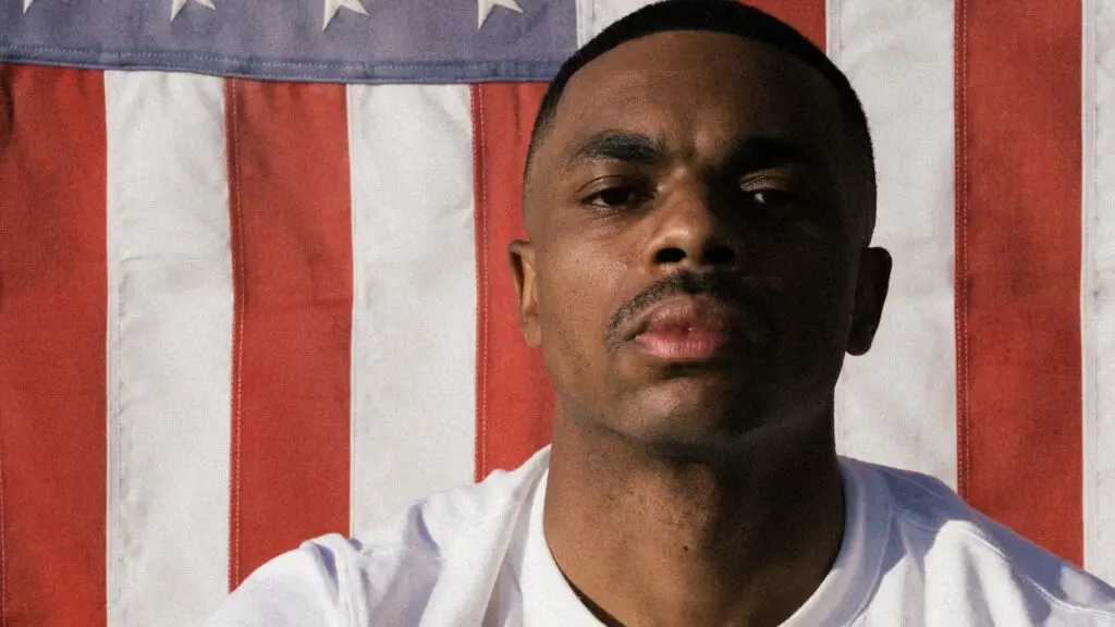Vince Staples press photo, close portrait against American flag backdrop