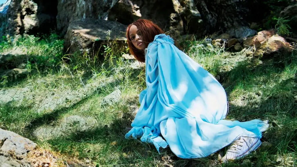 Ravyn Lenae in a flowing sky-blue gown on a rocky hillside, Blue Island era press photo