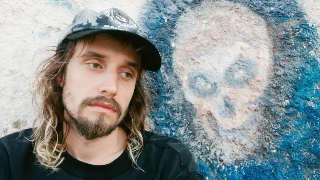 Pouya leaning against a graffiti wall with skull artwork, long hair and black cap, press photo
