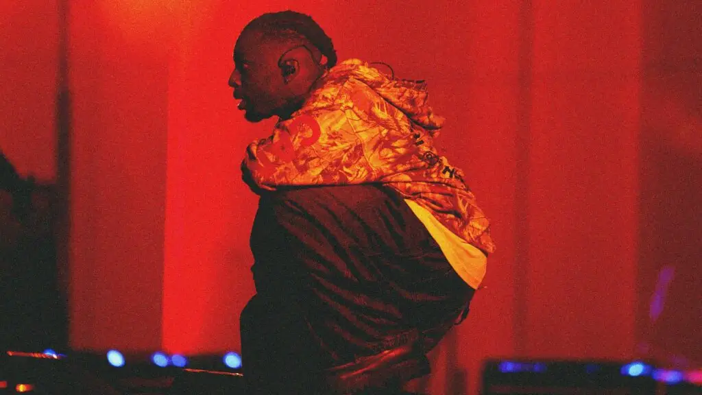 Isaiah Rashad performing live on stage, mid-show in camo hoodie under dramatic red lighting
