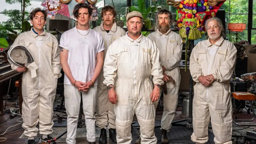 Modest Mouse band members in white jumpsuits in a studio setting with instruments and colorful decor
