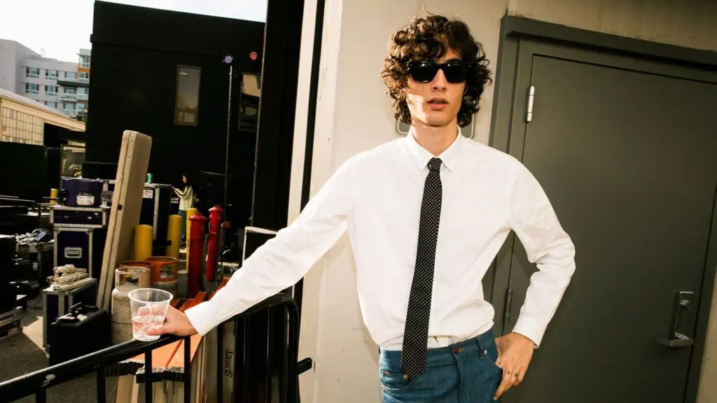 Sombr backstage in white dress shirt and tie with sunglasses, road cases behind him