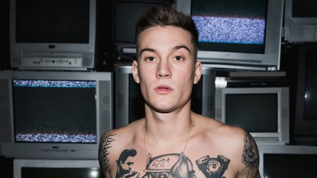 Cameron Whitcomb official press photo, shirtless with chest tattoos, against stacked vintage CRT televisions