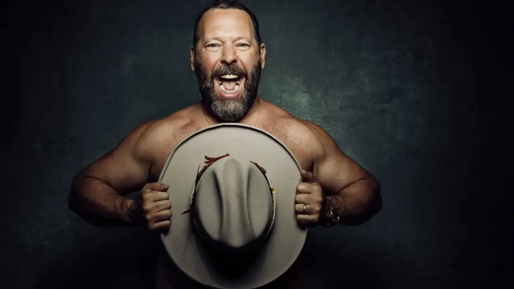 Bert Kreischer shirtless, laughing and holding a cowboy hat, official press photo