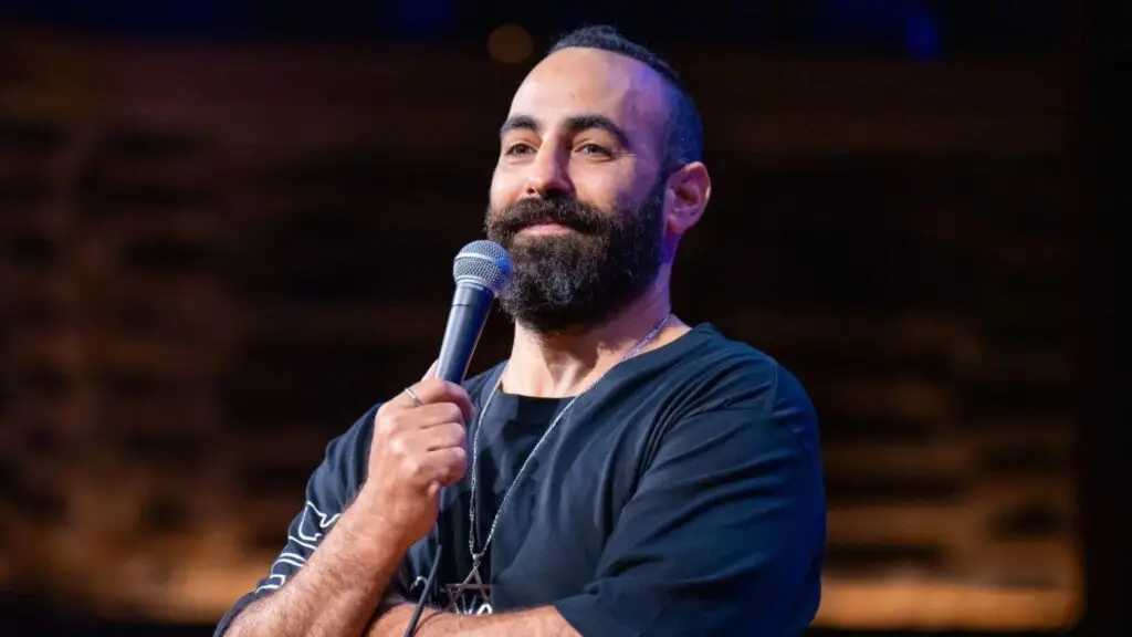 Yohay Sponder performing stand-up comedy on stage, holding microphone, Star of David necklace