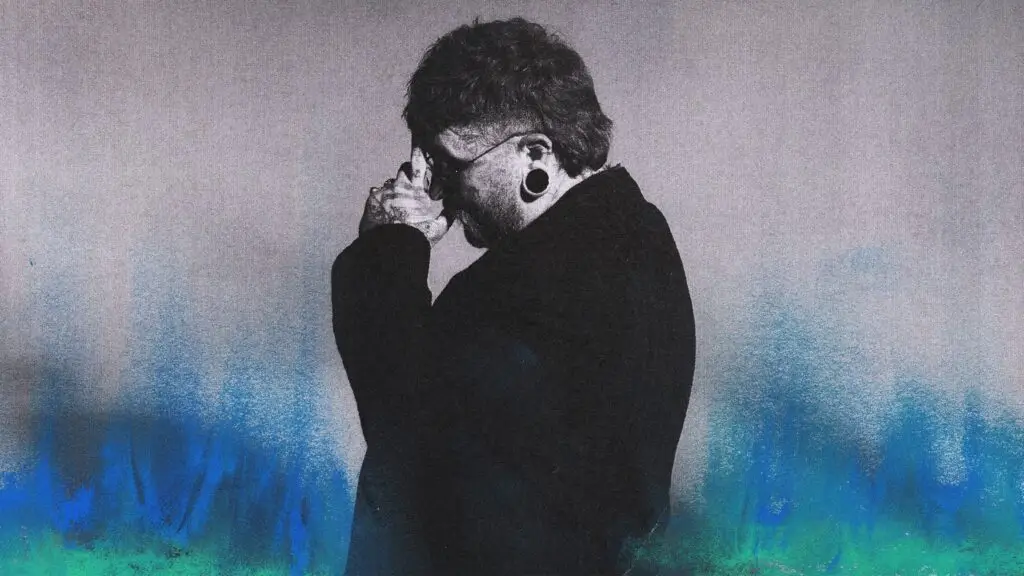 Teddy Swims press photo, head bowed with hands clasped, large ear gauges, black jacket, blue-green artistic accents