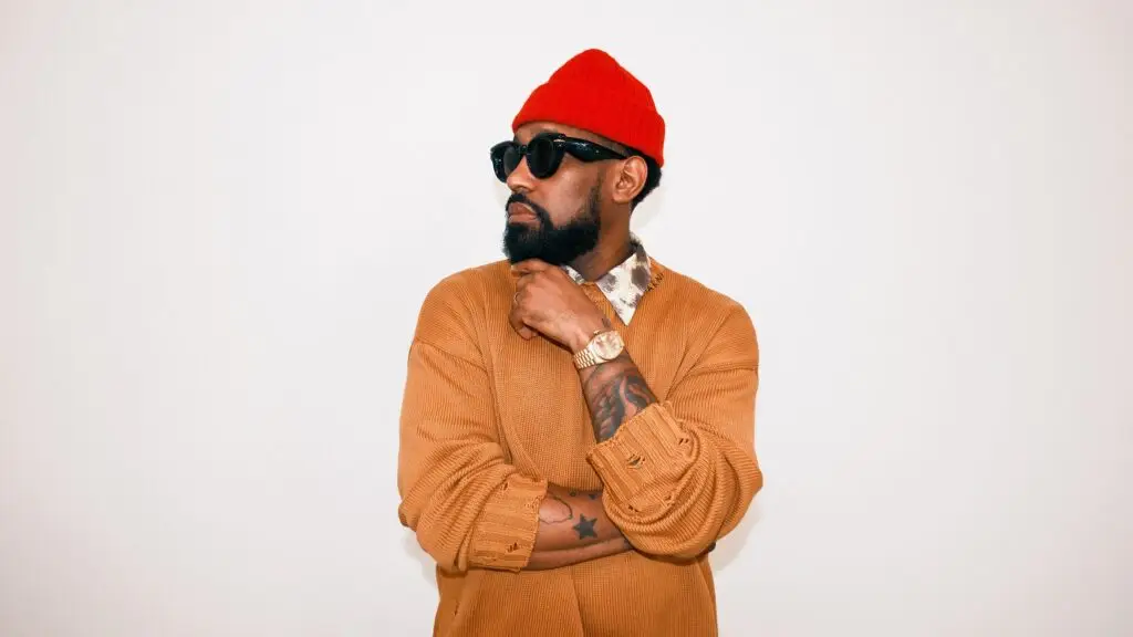 PJ Morton poses in a red beanie and tan cardigan for a studio press photo