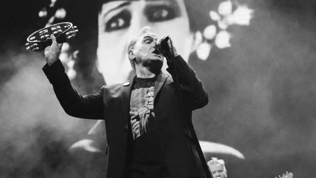 Morrissey performing live, tambourine raised, singing into microphone with face-projection backdrop
