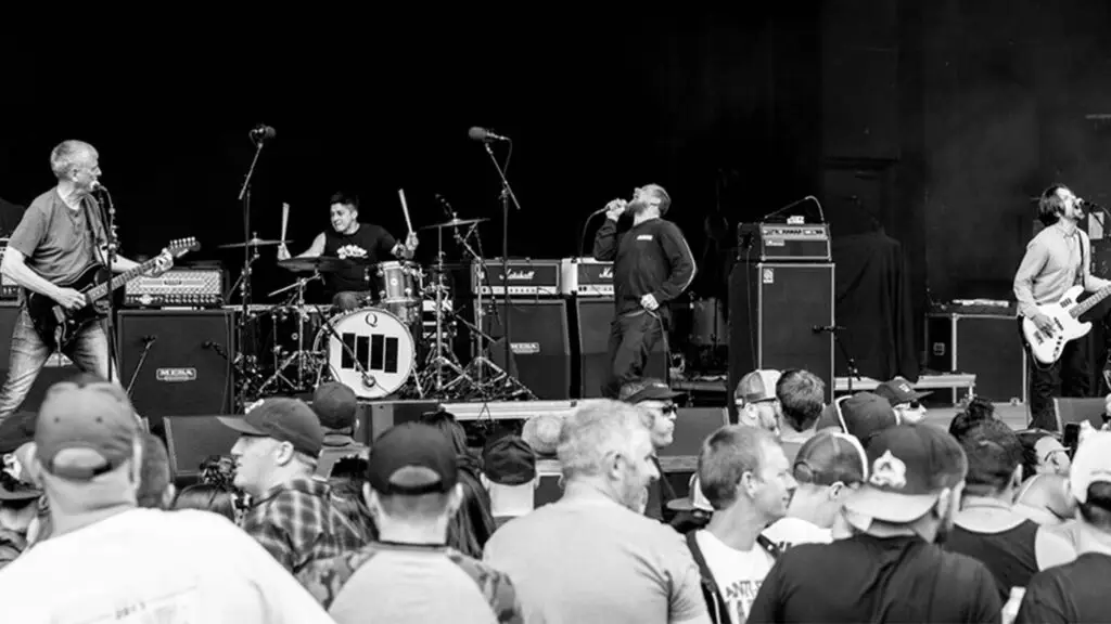 Black Flag performing live, Greg Ginn on guitar left, vocalist screaming center, crowd in foreground